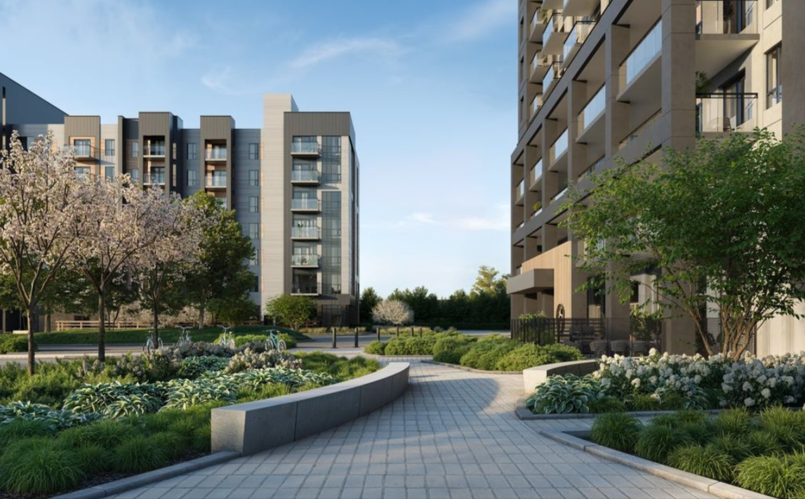 A paved walkway curves between landscaped gardens and modern apartment buildings under a clear blue sky.
