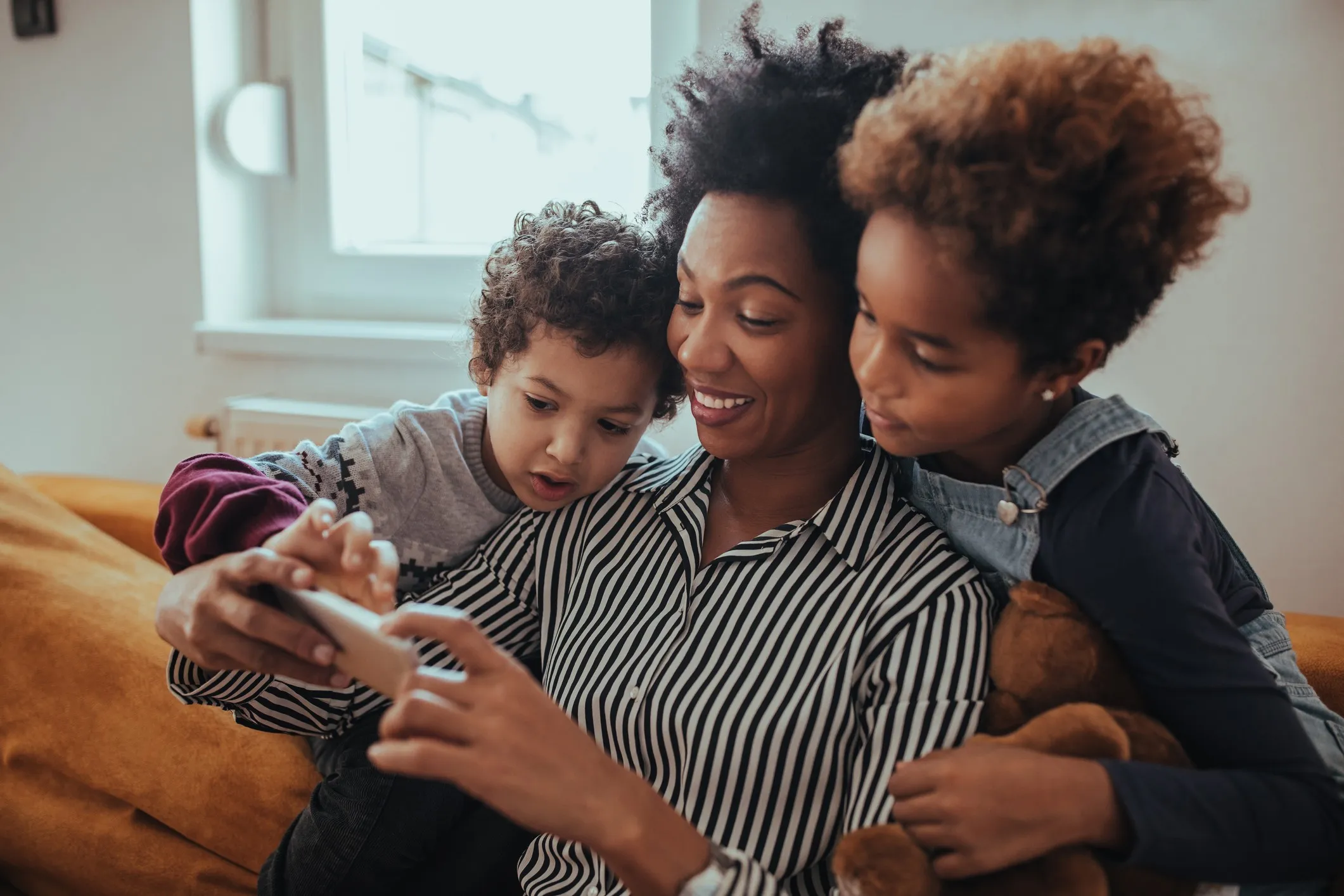 A mother and her two children sit on a couch, looking at a smartphone together, discussing ways to prevent child identity theft. 